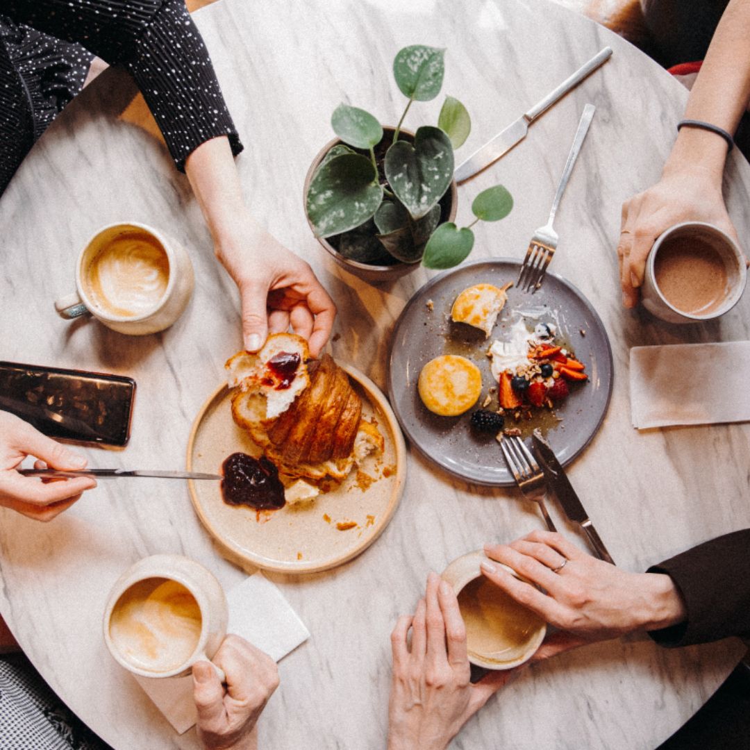 Image of plates of breakfast food, coffees, and hands all on a wooden table.